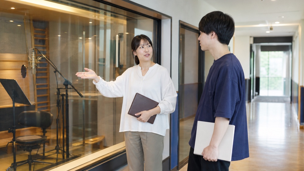 録音ブース見学中に学校スタッフへ質問する学生