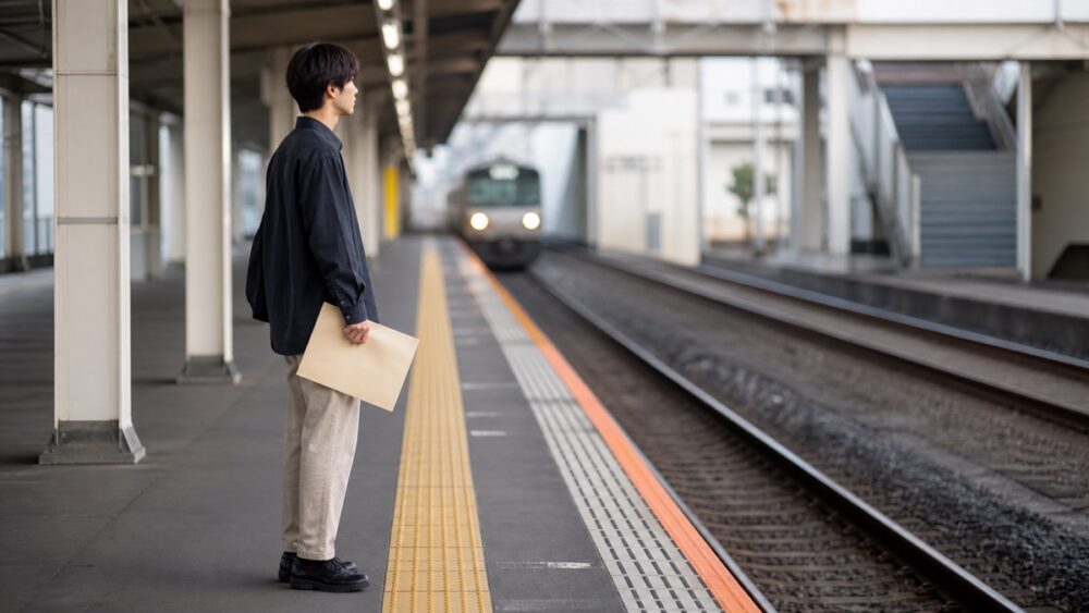 駅のホームで声優養成所へ向かう電車を待つ男性