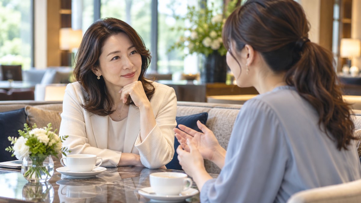 休日の昼、ホテルのラウンジの落ち着いたソファ席で、40代の女性が向かいに座る30代の女性の話をうなずきながら聞き、相談に乗っている近景。テーブルの上に文字は見えない