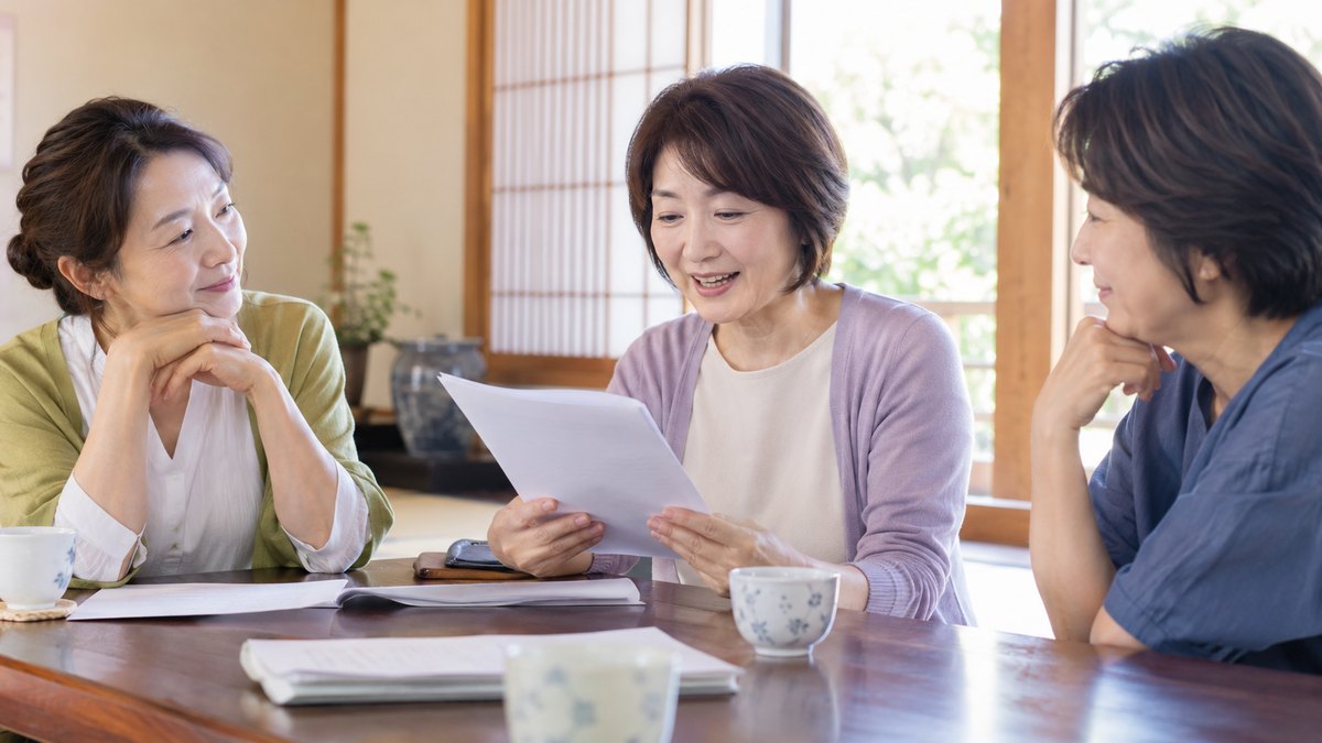 休日の朝、地域の公民館の明るい和室で、50代の女性3人が座卓を囲んで座り、1人が紙の原稿を声に出して読み、2人が穏やかに聞いている近景。原稿や壁の文字は見えない
