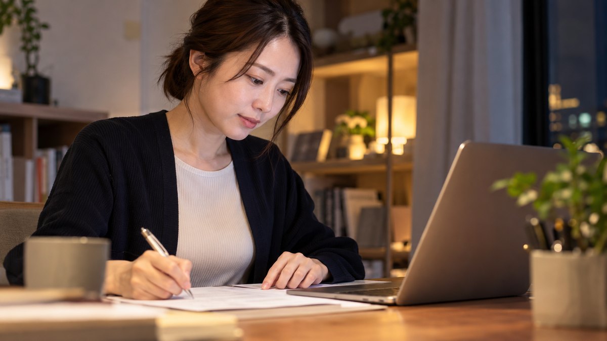 平日の夜、自宅の書斎の机で、30代の女性会社員がノートパソコンの横に職務経歴書のような紙を置き、ペンを持って自分の経験を書き出している近景。紙と画面の文字は読めない