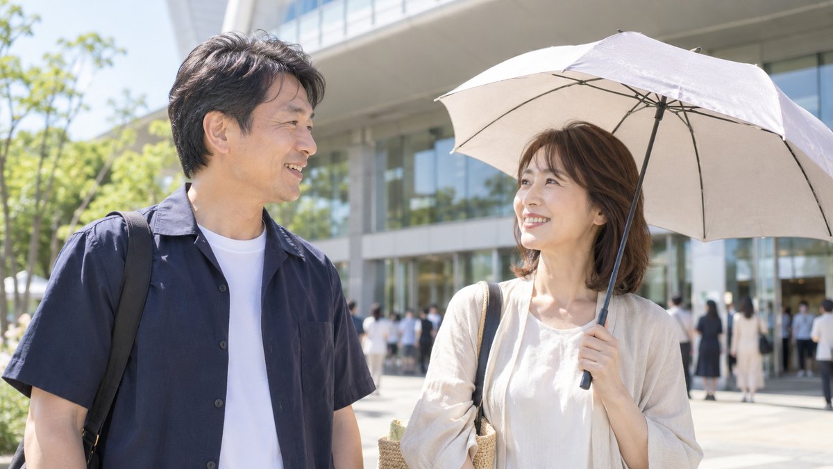 夏の朝、大きなホールの入口前の明るい広場で、40代の父親と日傘を持った40代の母親が並んで立ち、開場を待ちながら穏やかに話している近景。看板や建物の文字は見えない