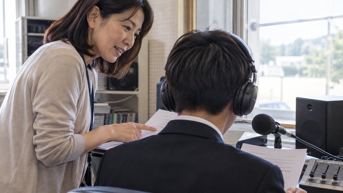 放課後、高校の小さな放送室で、ヘッドホンをつけてマイクの前に座る短髪の男子高校生の後ろ姿と、その横に立って原稿の一か所を指さしながら話しかける40代の女性教師の近景。教師の顔が見え、生徒は後ろ姿。原稿や機材の文字は見えない