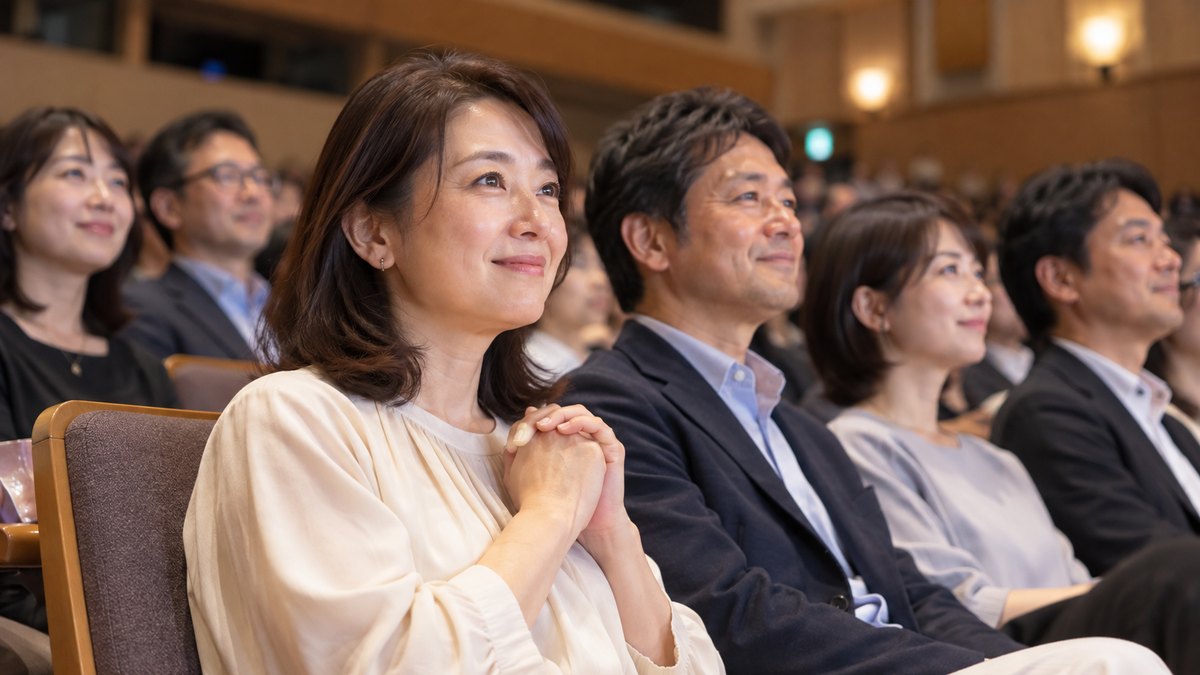 週末の午後、地方の市民ホールの客席で、30代〜50代の保護者たちが並んで座り、ステージのほうを静かに見守っている。手前の40代の女性が両手を胸の前で軽く組み、少し緊張しながらも温かい表情をしている。プログラムや壁の文字は見えない