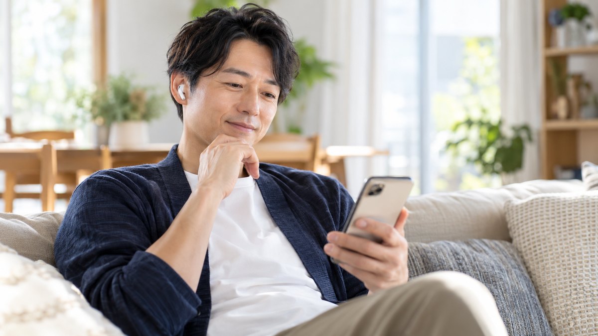 休日の午前、自宅の明るいリビングのソファで、40代の男性がイヤホンをつけ、手に持ったスマホで自分の話した声の録音を聞き返しながら小さくうなずいている近景。スマホの画面の文字は見えない