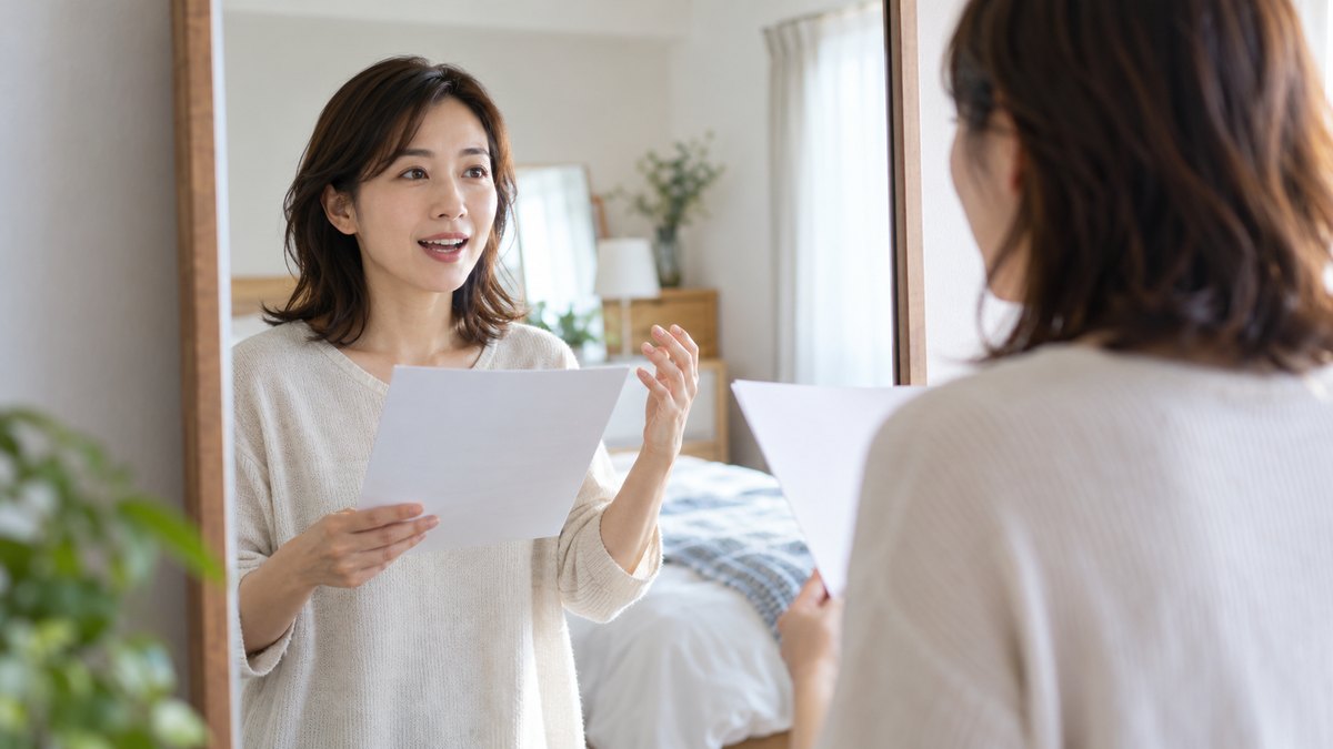 休日の朝、自宅の明るい寝室の姿見の前で、30代の女性が1枚の原稿を手に持ち、鏡の中の自分を見ながら声に出して読む練習をしている近景。原稿の文字は見えない