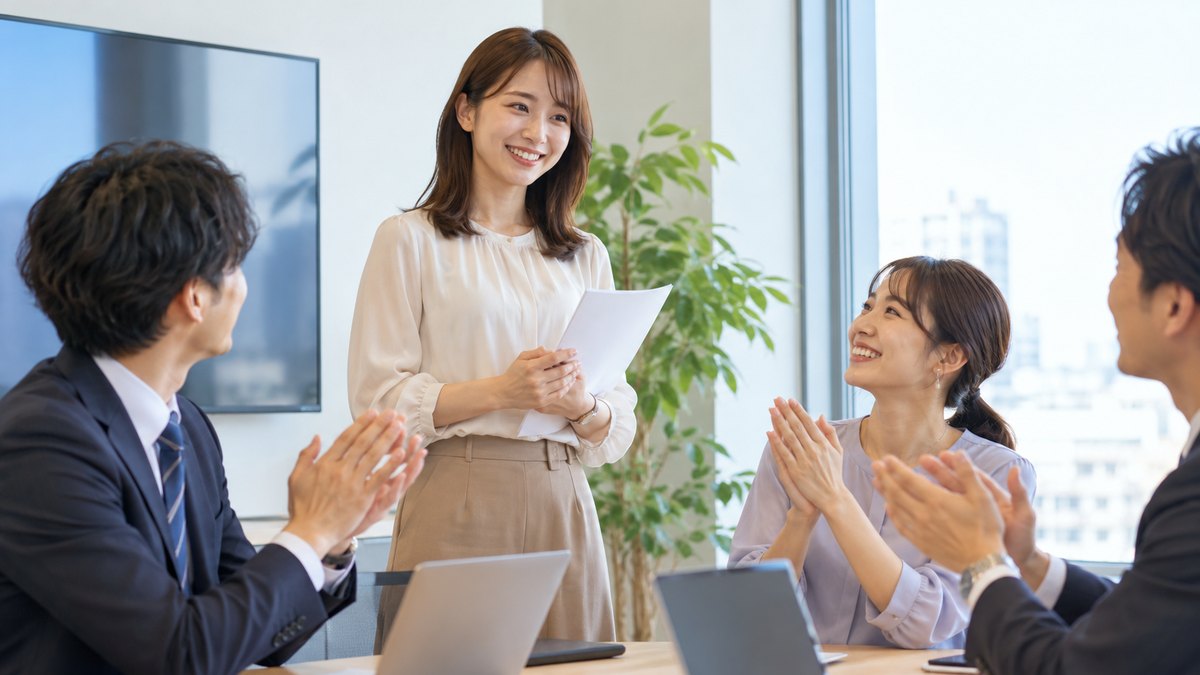 平日の午後、会社の小さな会議室で、30代の女性会社員が同僚3人の前で短い発表を終え、同僚たちが温かくほほえんで拍手している近景。モニターや資料に文字は見えない
