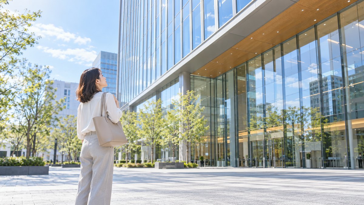 平日の昼、ガラス張りの大きな建物の前の広い広場で、30代の女性がトートバッグを肩にかけて立ち止まり、建物を見上げている遠景。建物や看板に文字やロゴは見えない