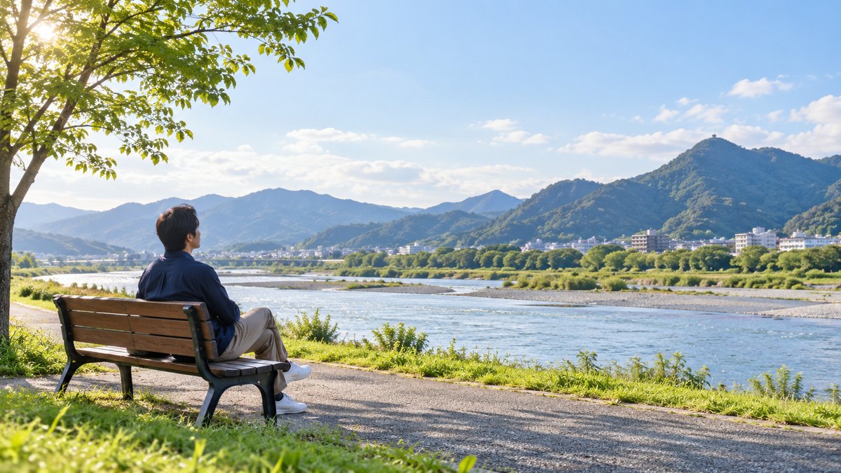 休日の朝、岐阜の大きな川の河川敷の遊歩道で、30代の男性がベンチに座り、川と山並みを眺めながら穏やかに深呼吸をしている遠景。看板や文字は見えない