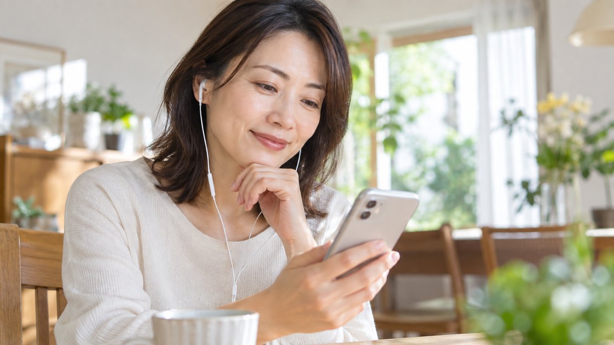 休日の午前、自宅の明るいダイニングで、40代の女性がイヤホンをつけ、手に持ったスマホで自分の話した声の録音を聞き返しながら小さくうなずいている近景。スマホの画面の文字は見えない