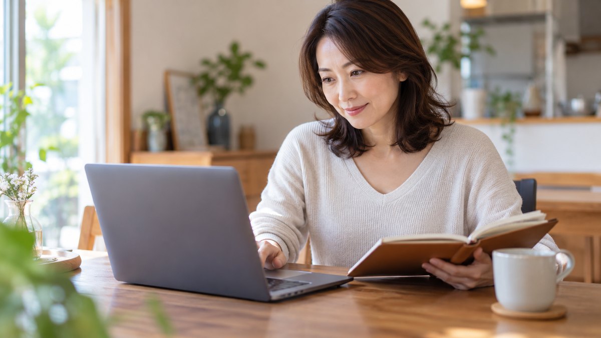 休日の午後、自宅のダイニングテーブルで、40代の女性がノートパソコンの前に座り、開いた手帳と見比べながら、問い合わせのメールを書いている近景。パソコンの画面と手帳の文字は見えない