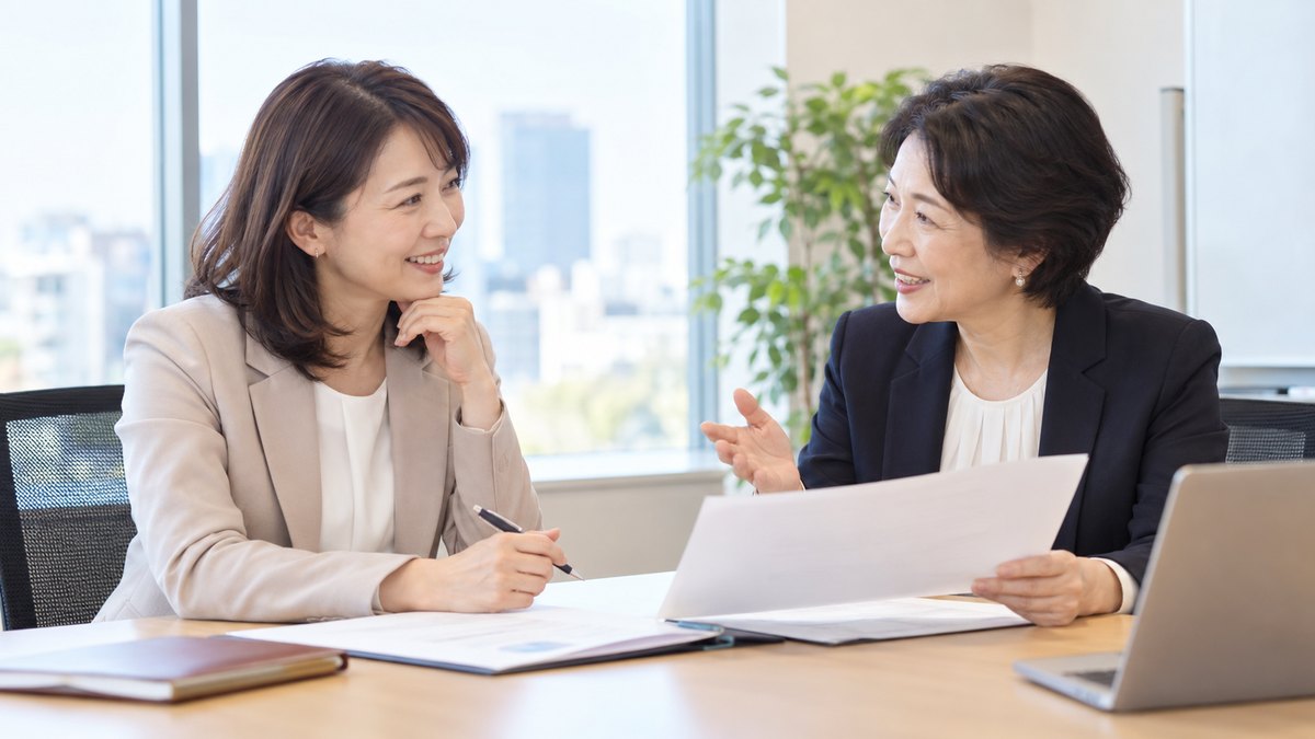 平日の夕方、明るいオフィスの小会議室で、40代の女性会社員が椅子に座り、向かいの50代の女性講師と書類を挟んで笑顔で相談している近景。書類の文字は見えない