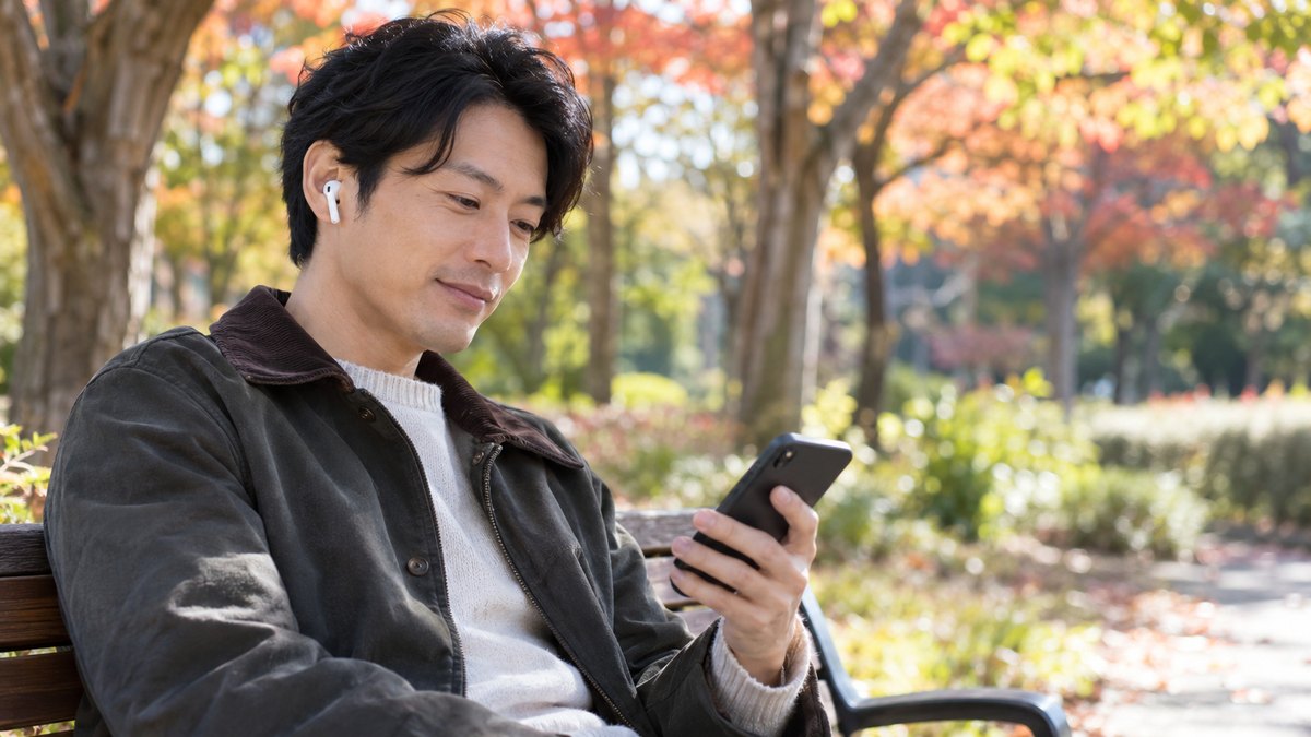休日の午前、紅葉の始まった公園のベンチに、30代の男性が座り、イヤホンをつけて手に持ったスマホで自分の話した声の録音を聞き返しながら小さくうなずいている近景。スマホの画面の文字は見えない