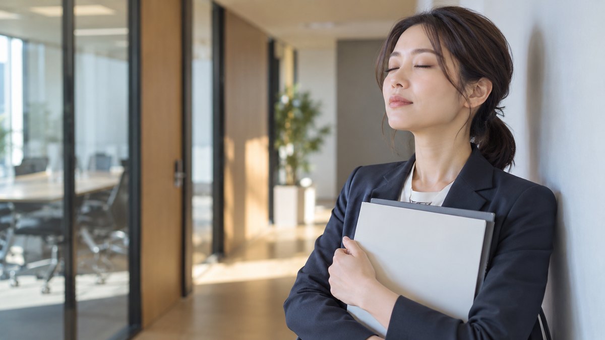 朝、会社の会議室の前の静かな廊下で、30代の女性会社員が壁ぎわに立ち、資料を胸に抱えて目を閉じ、ゆっくり息を吐いている近景。資料の文字は見えない