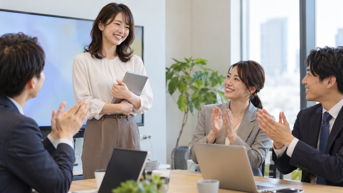 平日の午後、会社の小さな会議室で、30代の女性会社員が同僚3人の前で短い発表を終え、同僚たちが温かくほほえんで拍手している近景。モニターや資料に文字は見えない