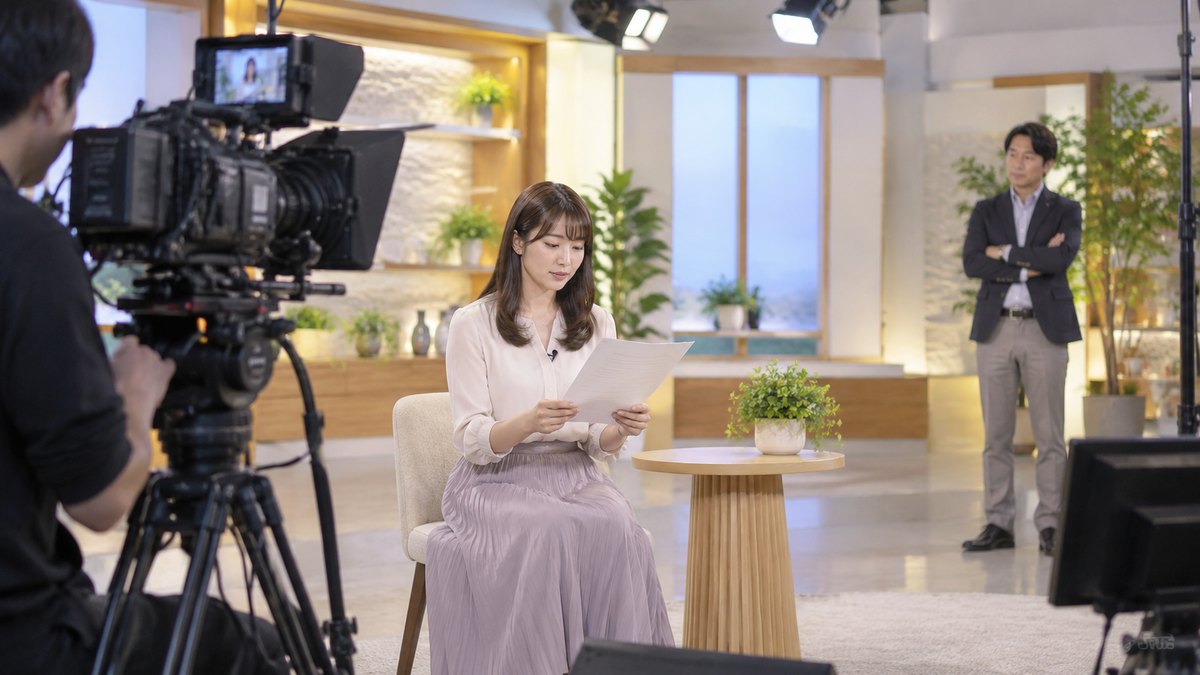 平日の昼、照明の当たった小さなテレビスタジオで、20代前半の女性がカメラの前の椅子に座って原稿を読み、少し離れた所で40代の男性講師が腕を組んで静かに聞いている遠景。カメラやセットに文字やロゴは無い
