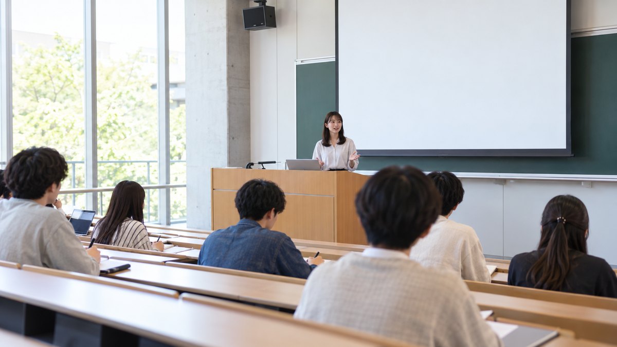 平日の昼、大学の階段教室の前方で、20代前半の女子大学生がゼミの発表をしていて、前の列に座る数人の学生がノートを取りながら聞いている遠景。スクリーンや黒板に文字は見えない