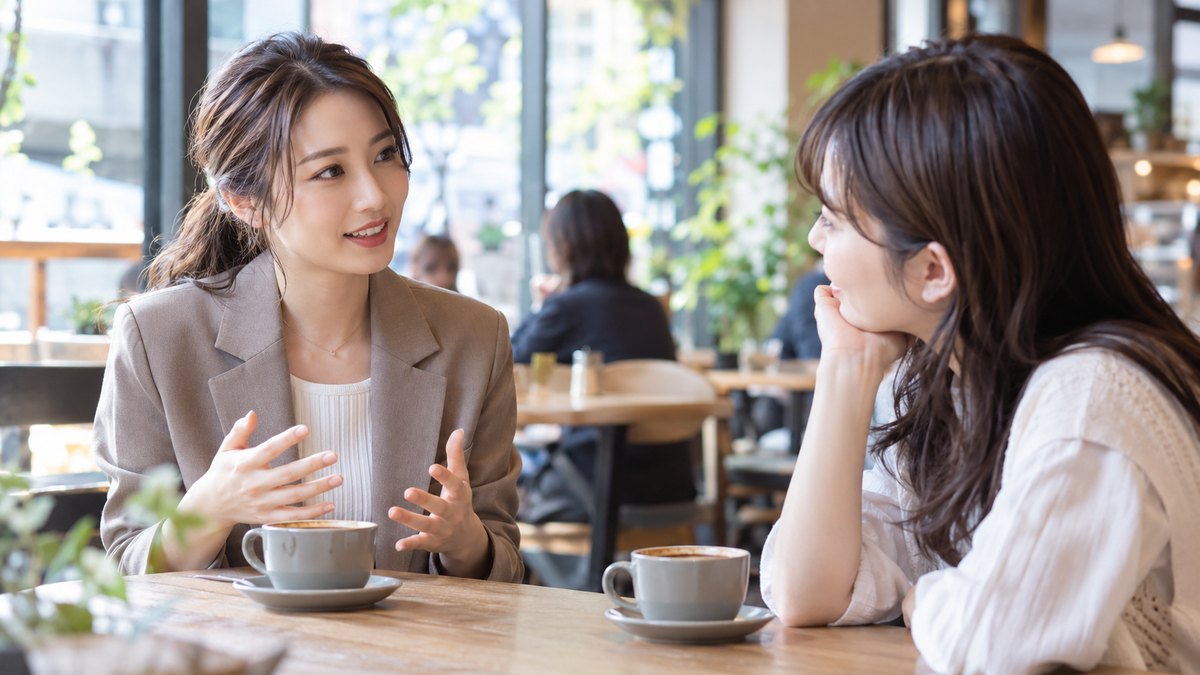 休日の午後、街のカフェのテーブル席で、20代後半の女性会社員が向かいに座る20代前半の女子大学生の話にうなずきながら、手ぶりをまじえて自分の就活の経験を話している近景。テーブルの上にコーヒーが2つ。文字は見えない