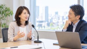 研修ルームで卓上マイクの前に座り、男性講師に話し方の助言を受けながら話す女性会社員