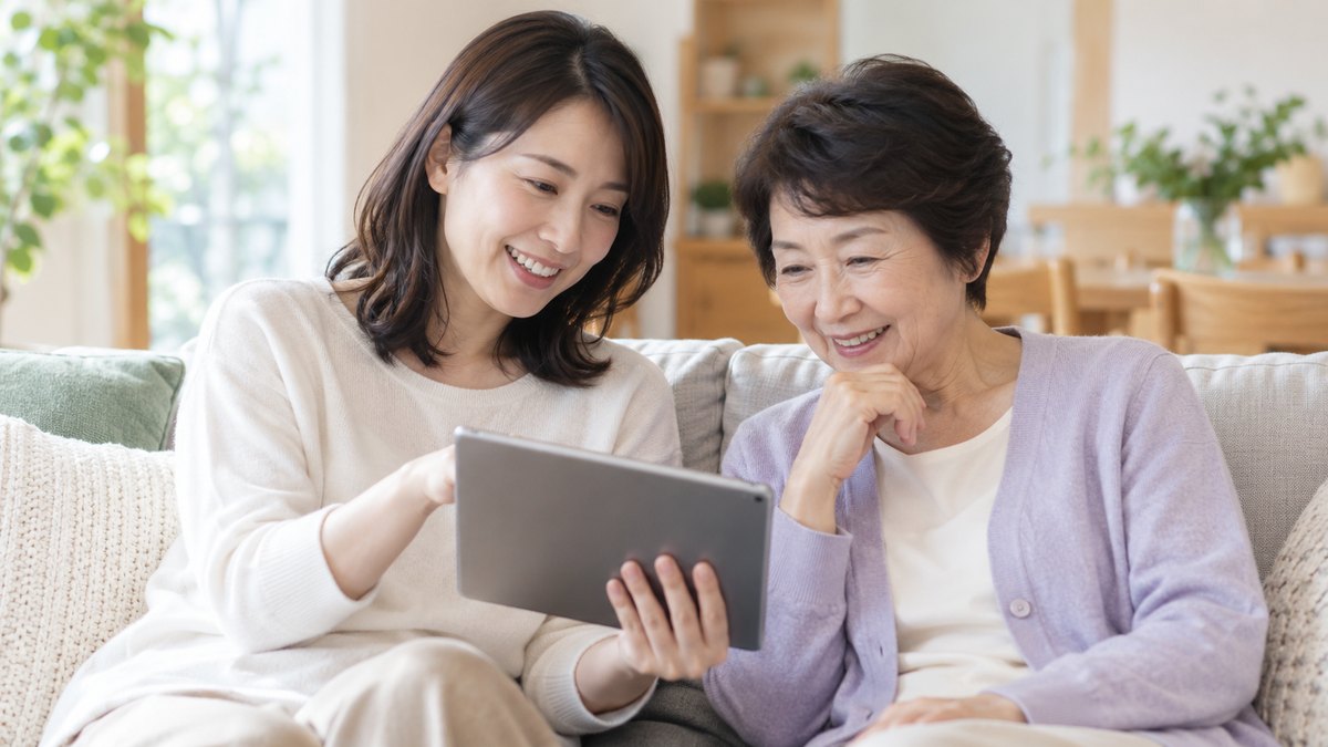 休日の午前、自宅のリビングのソファで、30代の女性がタブレットを膝にのせ、隣に座る60代の母親に画面を見せながら穏やかに話している近景。画面の文字は見えない