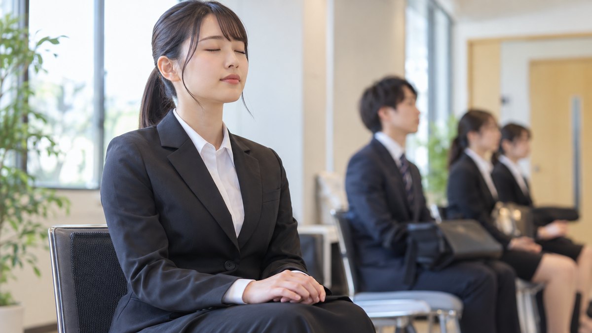 平日の朝、放送局の採用試験の控室で、スーツ姿の20代前半の女子大学生が椅子に座って膝の上で手を重ね、目を閉じてゆっくり息を吐いている近景。奥に順番を待つ学生がぼけて見える。掲示物や扉に文字は見えない