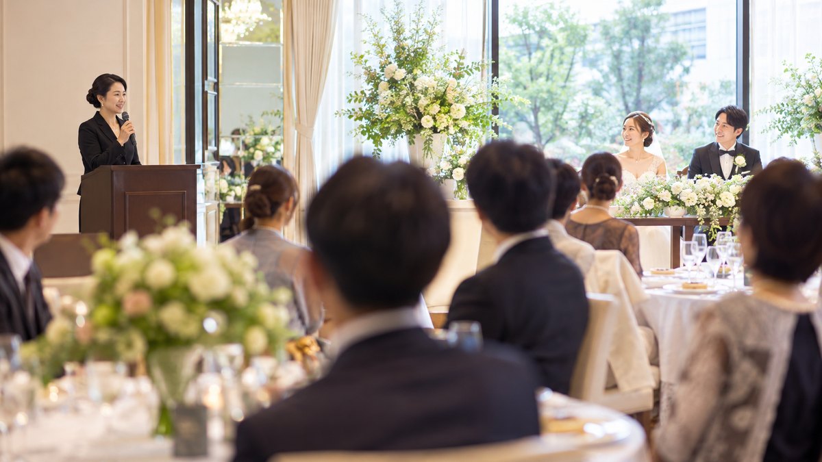 週末の昼、ホテルの明るい宴会場の結婚披露宴で、黒いスーツ姿の40代の女性司会者が会場の端の司会台に立ち、マイクを持って穏やかな笑顔で進行していて、手前の円卓に座る招待客がぼけて見える遠景。看板や席札の文字は見えない