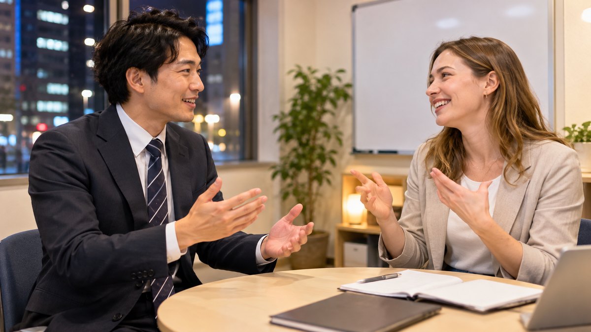 平日の夜、駅前の語学スクールの小さな教室で、スーツ姿の30代の男性会社員が丸テーブルをはさんで外国人の女性講師と英会話の練習をしていて、2人とも笑顔で身ぶりをまじえて話している近景。ホワイトボードや掲示物に文字は見えない