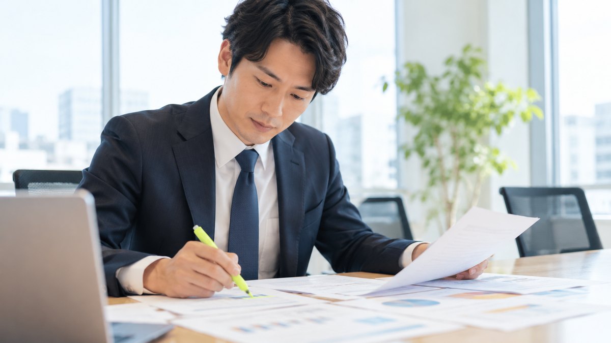 平日の昼、オフィスの明るい会議室で、30代の男性会社員が印刷した数枚の資料を机に並べ、蛍光ペンで線を引きながら見比べている近景。資料の文字は読めない