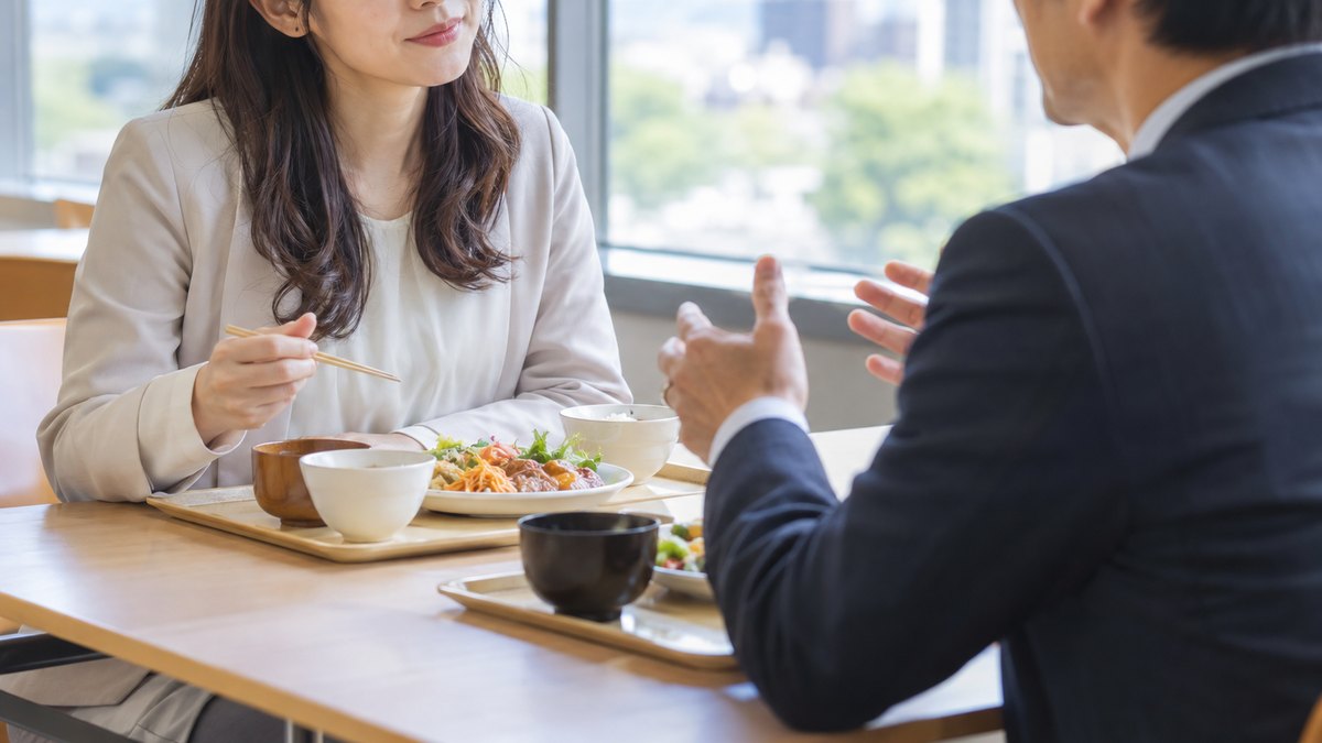 昼、社員食堂の窓際の席で、30代の女性会社員と40代の男性の同僚が向かい合ってトレーを前に座り、女性がうなずきながら相手の話を聞いている近景。顔ははっきり写らない。文字は無い