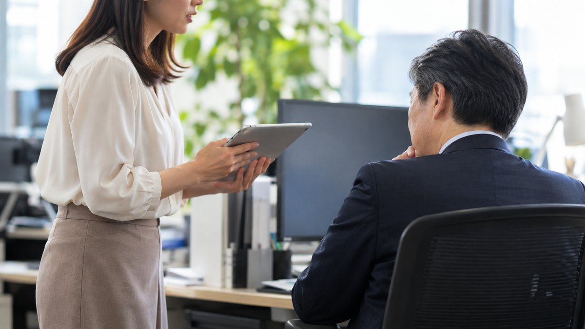 午後、オフィスの上司の席の横で、30代の女性会社員がタブレットを両手で持って立ち、椅子に座っている50代の男性上司に落ち着いて相談している横からの近景。顔ははっきり写らない。タブレットとモニターの画面は見えない角度。文字は無い