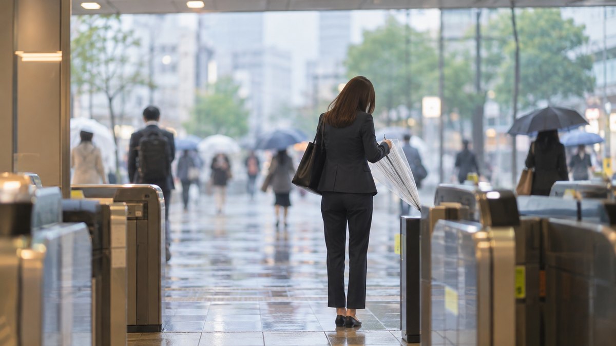 雨の日の朝、駅の改札を出た所で、30代の女性会社員が透明なビニール傘をたたんでいる後ろ姿の遠景。行き交う人はぼやけている。顔は写らない。看板や案内の文字は見えない