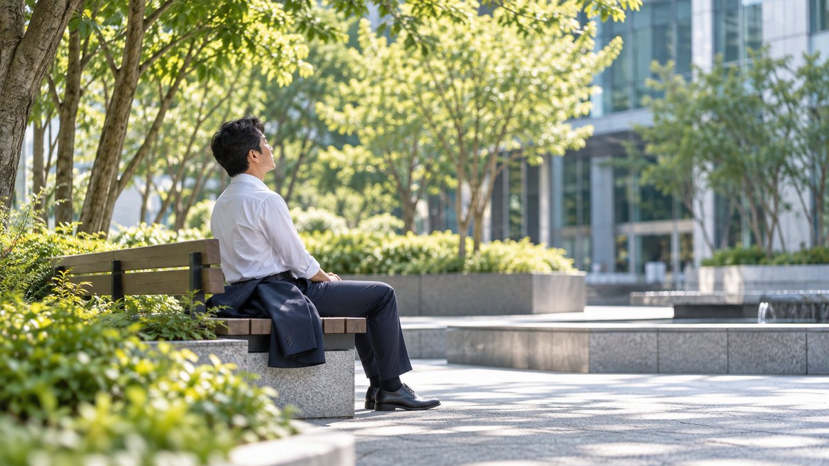 朝、オフィスビルの中庭の木陰のベンチで、スーツの上着を脇に置いた40代の男性会社員が、背すじを伸ばして目を閉じ、ゆっくり息を吐いている横からの遠景。顔ははっきり写らない。文字は無い
