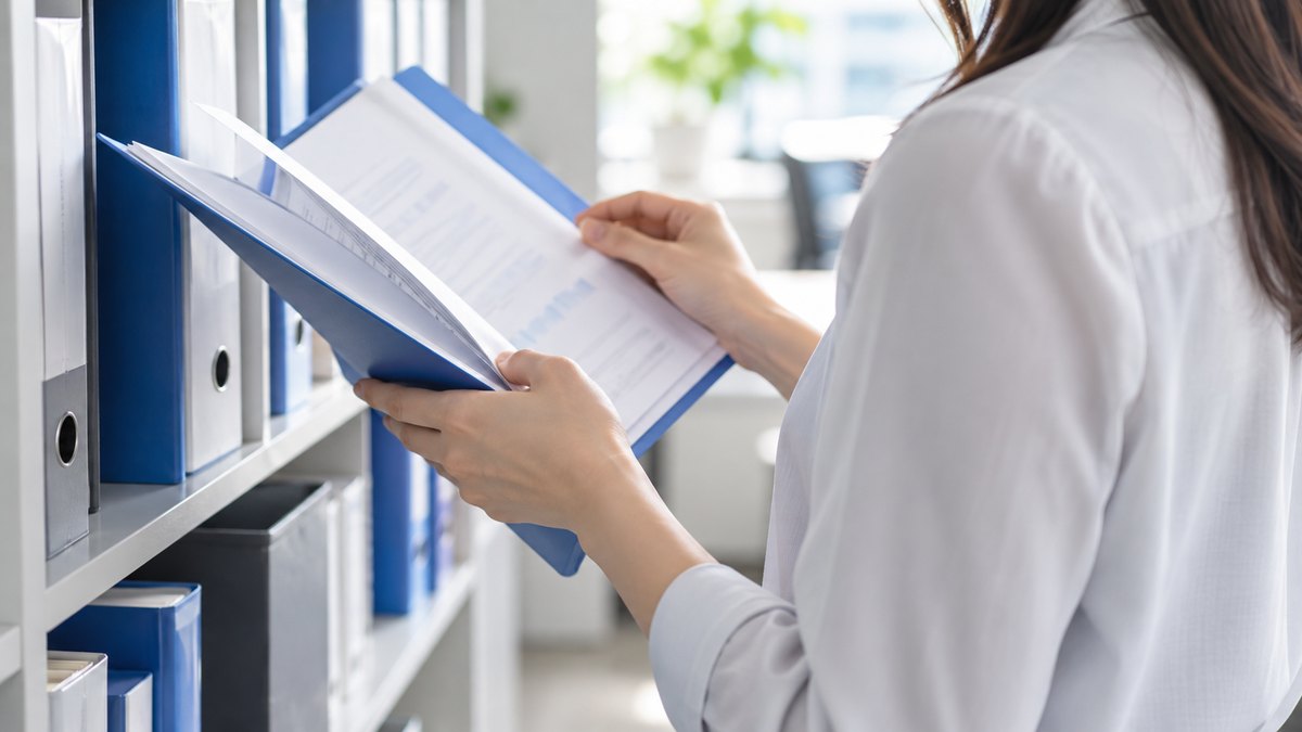 午前、オフィスの壁ぎわの書類棚の前で、30代の女性会社員が青いファイルを1冊引き出して中をめくっている手元と肩の近景。顔は写らない。ファイルと書類の文字は読めない