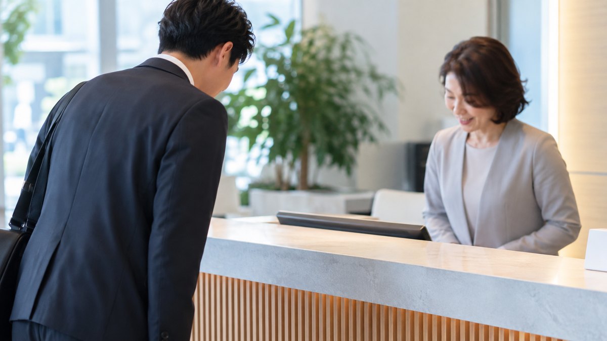 朝、オフィスの受付カウンターで、30代の男性会社員が受付の50代の女性に軽く頭を下げて、短くあいさつをしている近景。顔ははっきり写らない。受付の表示の文字は見えない