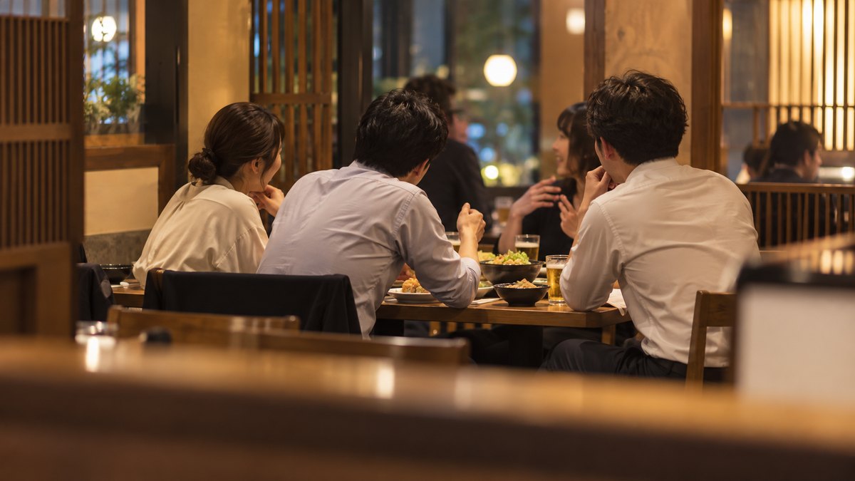 夜、会社の近くの居酒屋のテーブル席で、同僚4人が食事をしている中、30代の男性会社員が隣の人の話にうなずきながら短く質問している遠景。顔ははっきり写らない。メニューや店の文字は見えない