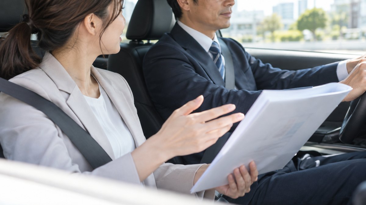 午前、営業車の助手席で、30代の女性会社員が運転する50代の男性の上司のほうを少し向いて、手元の資料を持ちながら話しかけている車内の近景。顔ははっきり写らない。資料の文字は読めない