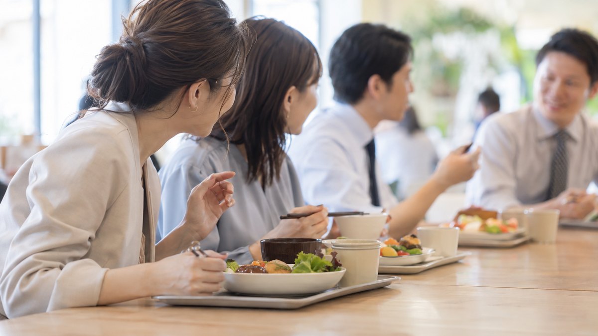 昼、社員食堂の長いテーブルで、トレーを前にした同僚4人が話している端の席から、30代の女性会社員が身を少し乗り出して、隣の同僚に短く質問している横からの近景。顔ははっきり写らない。メニューや掲示の文字は見えない