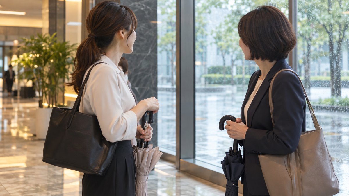 雨の日の朝、オフィスビルの1階のロビーで、傘をたたんだ30代の女性会社員が、同じく傘を持った40代の女性の同僚と窓の外の雨を見ながら軽く話している近景。顔ははっきり写らない。文字は無い