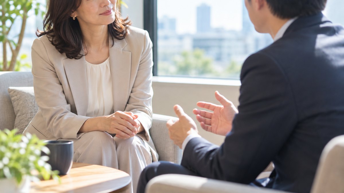 午後、明るいオフィスの窓ぎわの小さなソファ席で、40代の女性会社員と30代の男性の同僚が向かい合い、女性が相手の話に耳を傾けながら穏やかにうなずいている近景。顔ははっきり写らない。文字は無い