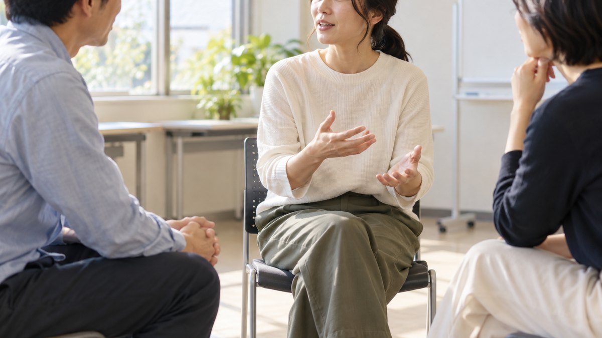 午後、明るい市民センターの小さな会議室で、30代から40代の大人3人が丸く座り、1人が短く話すのを2人が穏やかにうなずいて聞いている近景。顔ははっきり写らない。ホワイトボードや紙の文字は見えない