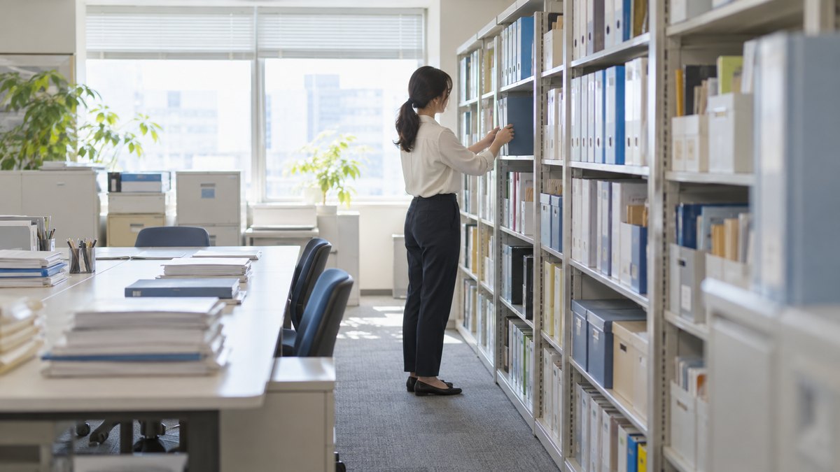 午前、会社の明るい資料室で、30代の女性会社員が棚からファイルを1冊取り出しながら、ひとりで静かに作業している横からの遠景。顔ははっきり写らない。ファイルや棚の文字は読めない