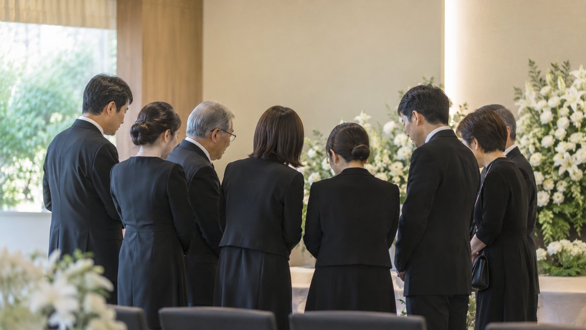 昼、小さな家族葬のホールで、喪服の家族8人ほどが前方の白い花のそばに集まり、低い声で話している後ろ姿。祭壇・遺影・棺は写らない