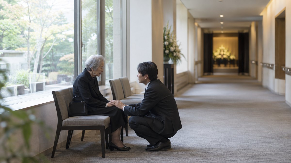 昼、斎場の廊下の窓辺で、喪服の80代の女性が椅子に座り、隣にしゃがんだ40代の孫の男性が、女性の手に自分の手を重ねて静かに話を聞いている。顔は小さく写る