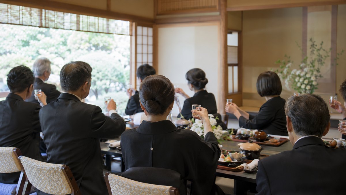 昼、料亭の座敷の長い座卓で、喪服の親族10人ほどが小さなグラスを胸の高さに静かに持ち上げている手元と後ろ姿。顔は写らない。文字は無い