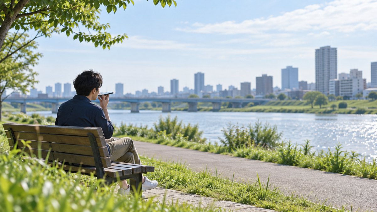 休日の朝、川沿いの河川敷のベンチで、30代の男性がスマホを口元に近づけて、小さな声で1分間話して録音している遠景。スマホの画面は見えない。顔ははっきり写らない。文字は無い