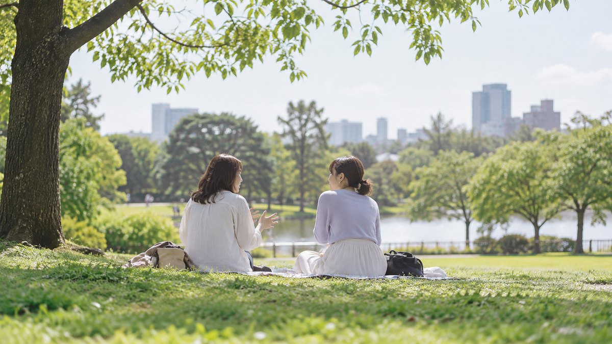 休日の昼、公園の芝生にレジャーシートを敷いて、30代の女性が同年代の友人と並んで座り、友人に悩みを話してうなずいてもらっている遠景。顔ははっきり写らない。文字は無い