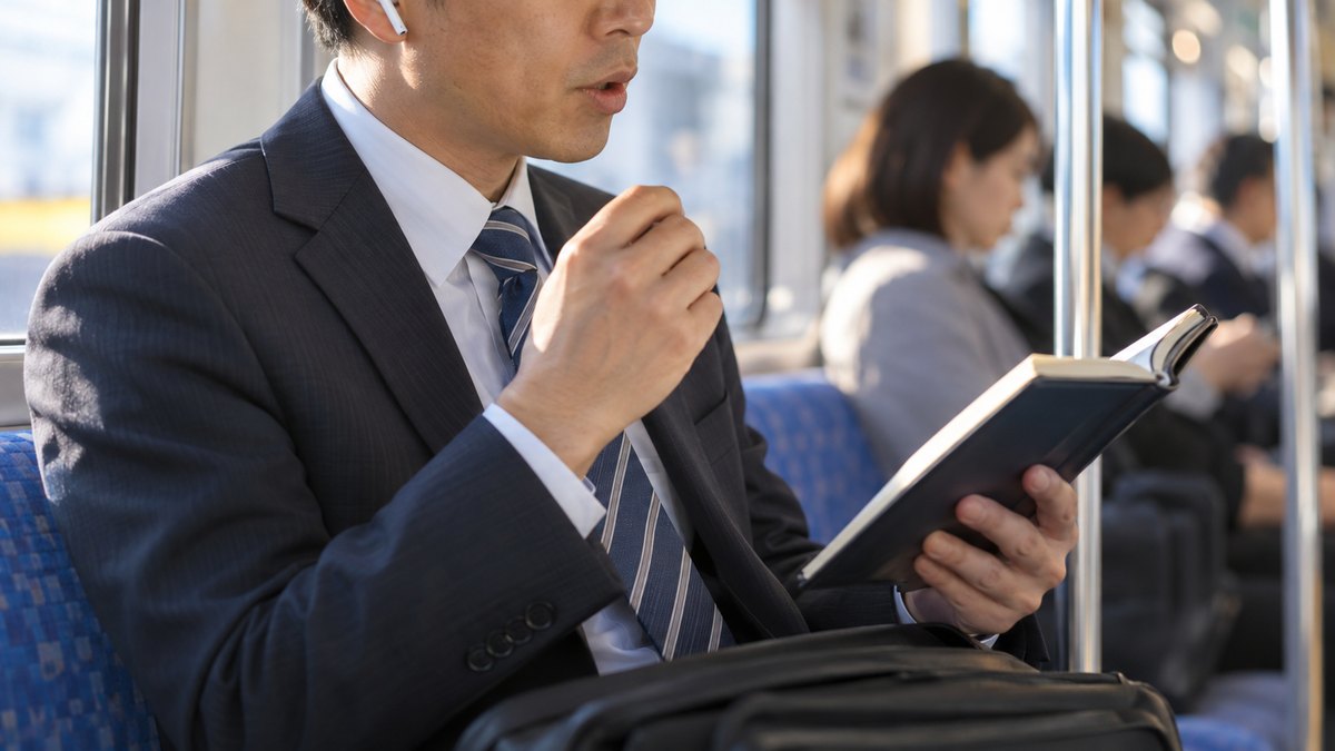 朝、通勤電車の座席で、40代の男性会社員がイヤホンをつけ、手帳に書いた短いメモを見ながら口元を小さく動かして話す練習をしている近景。手帳の文字は読めない。顔ははっきり写らない。文字は無い
