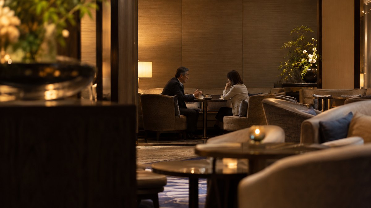 午後、ホテルのラウンジの窓のない奥の席で、50代の男性管理職が40代の女性コーチとコーヒーを前に向かい合い、身を乗り出して1対1で話している遠景。高層ビルは写さない。顔ははっきり写らない。文字は無い