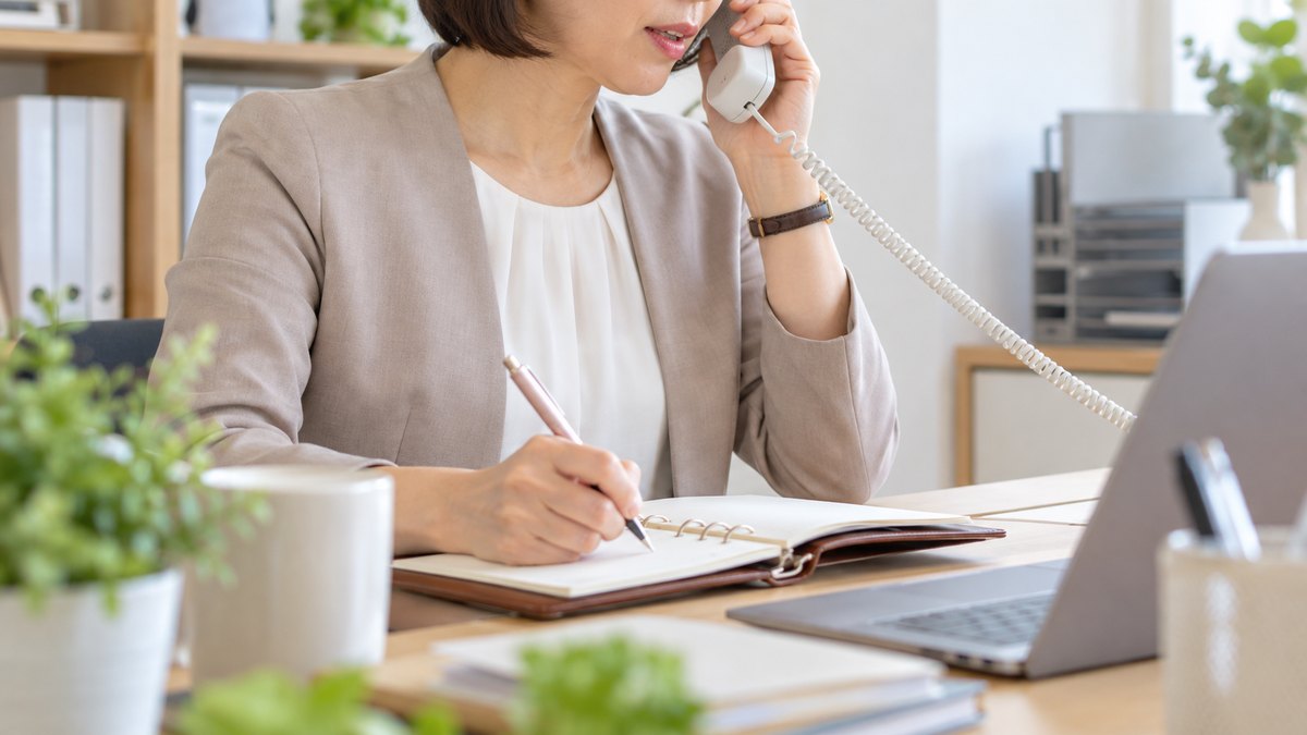 午前、研修会社の小さな事務所の自席で、40代の女性講師が電話の受話器を耳に当て、企業の人事担当と打ち合わせをしながら手帳に書き込んでいる近景。手帳と棚の文字は見えない。窓の景色は写さない。顔ははっきり写らない。文字は無い