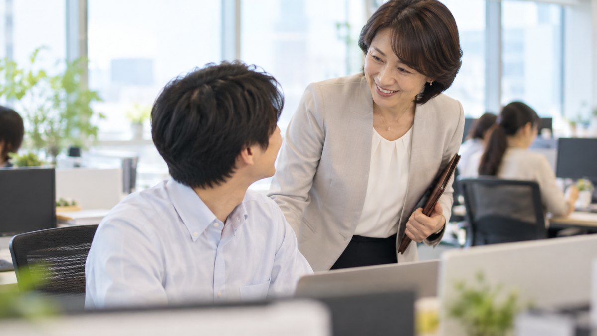 朝、オフィスの島型の席で、パソコンに向かう20代の男性社員のそばに50代の女性課長が立ち寄り、笑顔で声をかけ、社員が振り向いてうれしそうに会釈している近景。パソコンの画面は見えない。顔ははっきり写らない。文字は無い